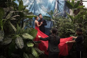 Assistants hold a bride’s red dress as she poses together with her groom