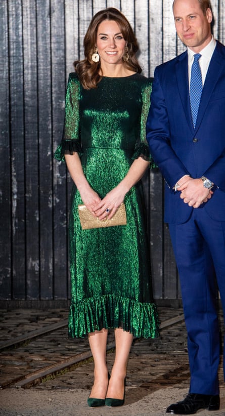 Kate seen full length in a long dark green sequined version of the dress it has ruffles at the elbows and hem, and she is wearing matching high heels and holding a small purse; golden hand. Her long black hair is down and wavy, and she wears large gold earrings. Prince William is partly seen next to her, wearing a dark blue suit, white shirt and tie; they stand against what appears to be a wall or partition made of thin, vertical black wooden planks. 