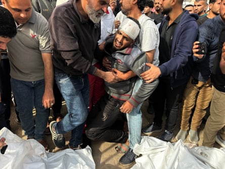 Men stand over bodies wrapped in white shrouds, with two men supporting a distressed man with a bandaged head