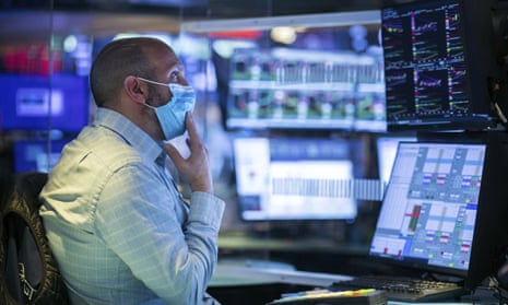 A trader in shirtsleeves and a face mask sits at a desk with several display screens in front of him