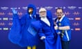 Man in outlandish blue suit and white short with blond hair with someone dressed in blue as a bird and a man in a blue top with an EU flag top either side of him.