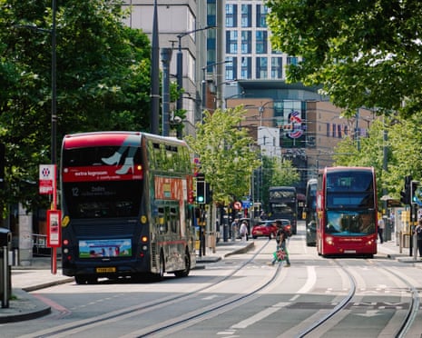 Two double-decker buses travel along a tree-lined Birmingham street with tram tracks and modern buildings behind