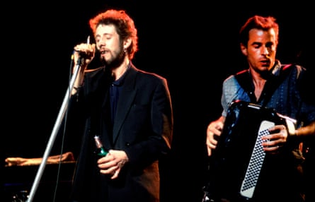 The perform on stage against a black background, MacGowan singing, Fearnley on accordion