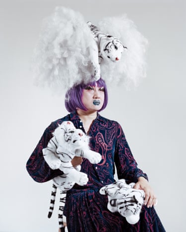 A person with purple hair and a huge white fluffy headpiece holds plush white tiger toys