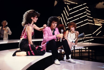 Leo Sayer sits on a stage with two women, wearing a pink blazer.