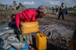 A woman pours water into a jerrycan, surrounded by the remains of her shelter as people pass by