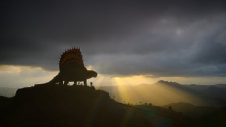 The silhouette of a dinosaur is seen on the crest of a hill with a dramatic sunburst in the distance