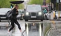 A woman holding an upturned umbrella leaps over a large puddle on a road in London as a taxi approaches in the distance