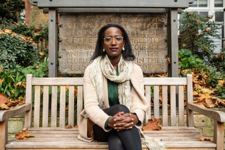 A young woman sits cross-legged on a bench in London before a plaque looking at the camera