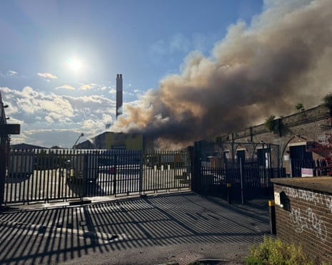 Smoke from a recycling plant seen behind a metal industrial fence rises into a blue sky against the sun