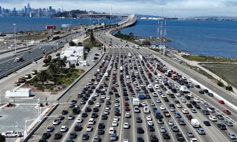 A wide freeway with 17 lanes is filled with vehicles heading toward the toll plaza on California's Bay Bridge. In the distance is the San Francisco skyline.