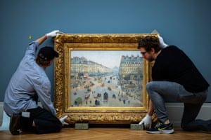 Two men wearing white gloves lift a painting with a gold frame off the floor
