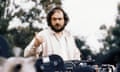 Kubrick looks toward camera and gestures over the top of a film camera