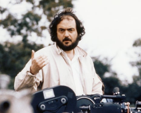 Kubrick looks toward camera and gestures over the top of a film camera
