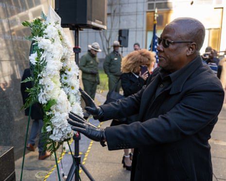 John Mahama, dressed in a black coat, black shirt, black leather gloves and sunglasses, puts his hands to a wreath of white flowers at a stone monument.