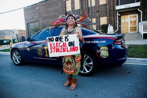 A woman in traditional Mexican clothes with feathers in her hair holds the hand-written sign