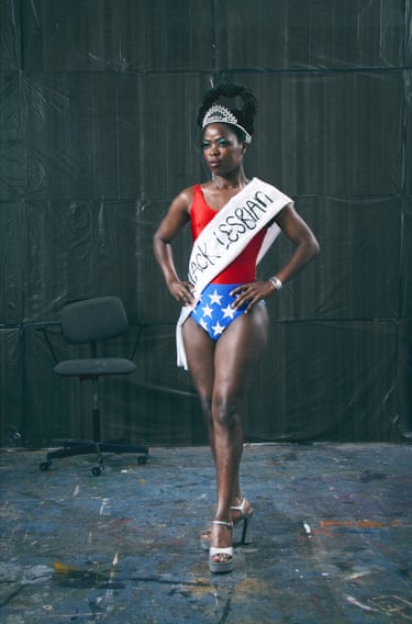 a woman in red, white and blue bodysuit with tiara and sash saying ‘Black lesbian’ stands with hands on hips