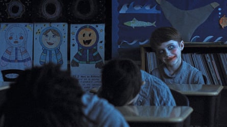 A young boy sits at classroom desk in dimly lit room wearing clown makeup