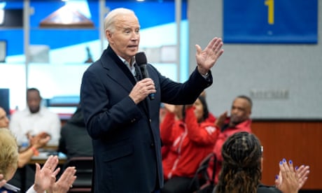 White man wearing blue peacoat speaks into microphone as people around him sitting at tables clap