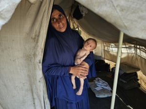 A woman wearing a blue khimar holds a malnourished baby inside a tent