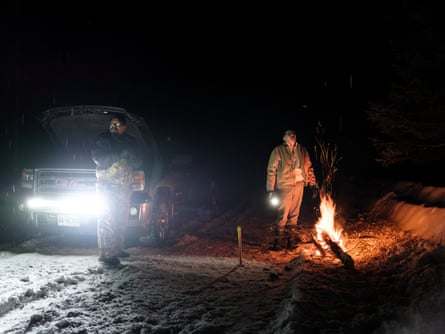 Two men in heavy overalls and boots stand in snow with a small fire of wooden branches to one side and the parked truck on the other, in the light of its headlamps.