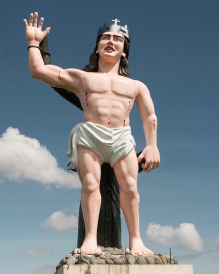 A model statue of a Christ-like figure wearing a crown with a cross on it is tied to a large cactus.
