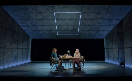 Two actresses sit at a table in a Some Woman stage scene, lit by a glowing square above