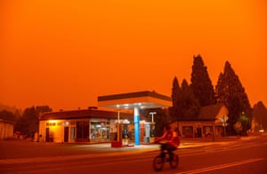 A man rides his bike past a gas station on 23 July as nearby flames turn the sky orange. The Dixie fire would destroy much of the town just days later.