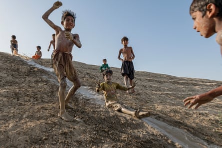 Gleeful mud-covered children stand on a slope, while one child slides down a water-covered channel on the slope