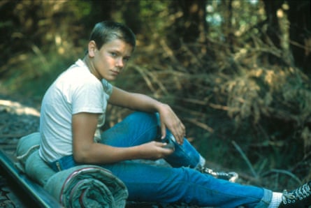 A young boy in a white T-shirt and jeans with short blond hair sitting on a sleeping bag in the forest