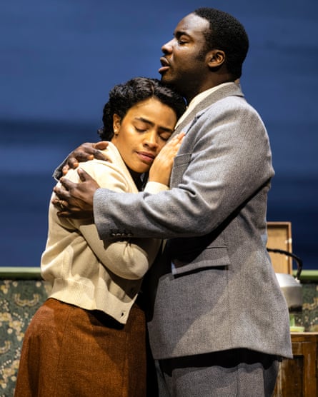 A black man in a grey suit and a black woman in a beige cardigan hug.
