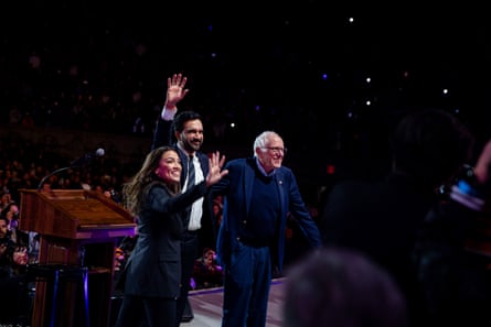 a woman and two men smile and wave on stage to crowd