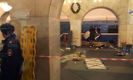 A man stands in an underground station near a blue train that is showing signs of explosive damage