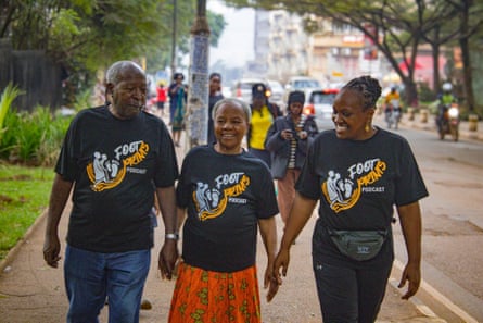 A woman and a older couple walk along a street. They are all wearing T-shirts with a Foot Prints podcast logo