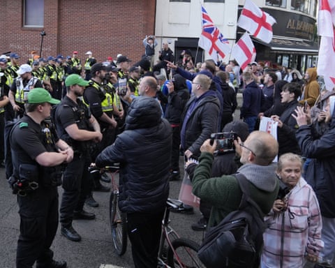 Demonstrators waving UK and England flags angrily confront a line of police officers.