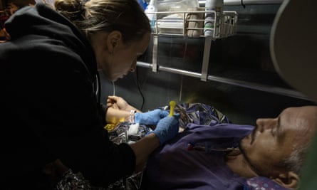 A female paramedic with blond hair tied into a ponytail leans over and injects a man wearing a blue sweatshirt who lies under a foil blanket on a bed in a converted medical bus