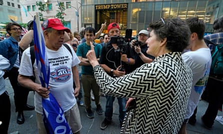 Middle-aged man in red hat holding blue flag faces middle-aged woman with brown hair and black and white blouse pointing her finger in his face.
