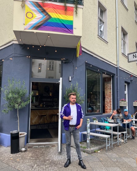 Danjel Zarte stands in the doorway of his cafe on a street corner; a Pride flag hangs above the door; people are sitting on tables in the street behind him.