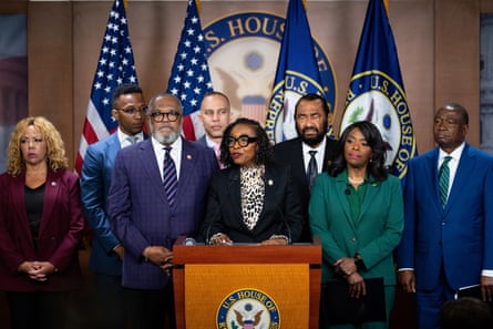A woman speaks at a lectern surrounded by people and in front of several flags.
