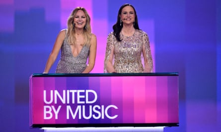 Hosts Malin Åkerman (L) and Petra Mede stand behind a piece of set furniture with 'United by Music' displayed on the front