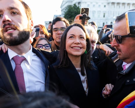 Wearing a dark jacket and white top, MarÃÂa Corina Machado smiles while surrounded by supporters, press and security during a visit to Washington DC