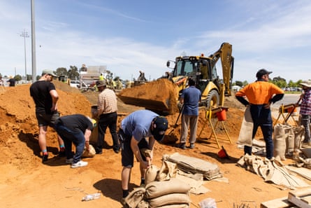 People are filling sandbags and shovelling sand from large piles of orange sand, and there is a digger in the background