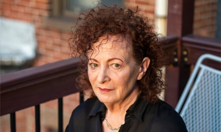 An older white woman with dyed red hair sits in the rays of the setting sun on a chair on a back stairway of an apartment building, looking seriously at the camera.