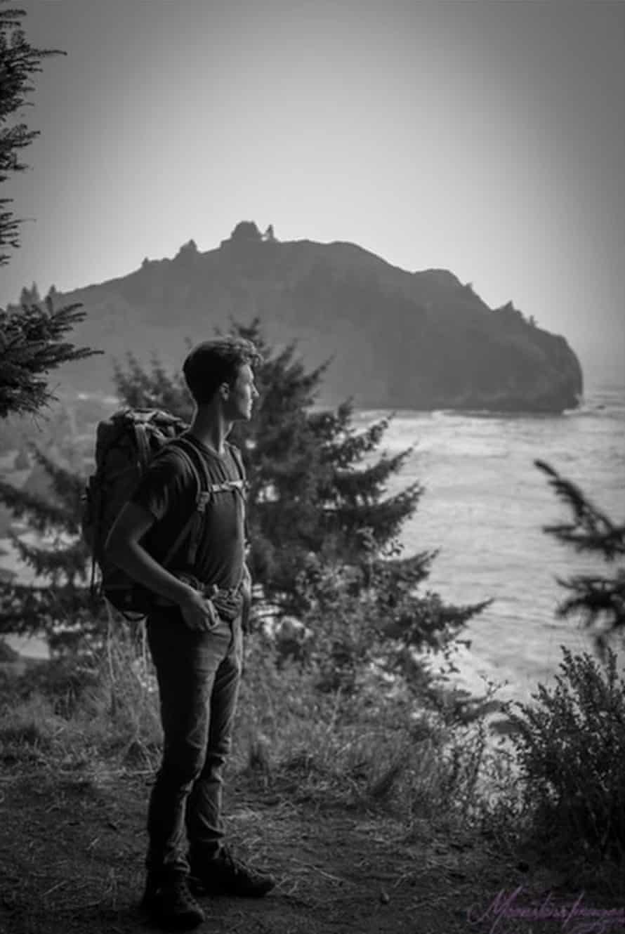A black and white portrait of a young man wearing a hiking backpack in a forested area while looking out to the sea. An island can be seen in the background.