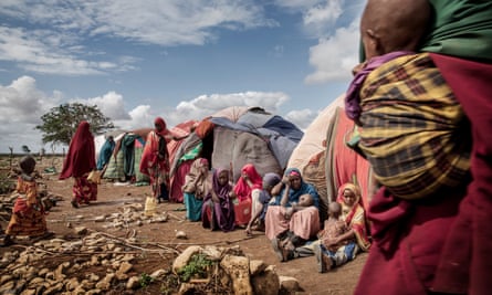 Women with babies and girls sit outside some basic shelters in a desert-like landscape