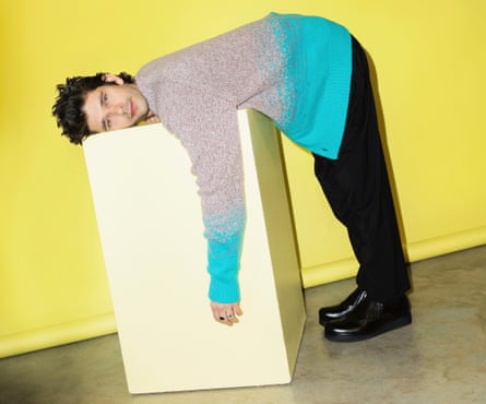 Actor Ben Whishaw in pink and blue cardigan and black trousers, leaning over a large white block, against yellow background, January 2022