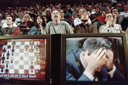 View of audience at chess tournament, with two monitors in foreground: one shows a chess board, the other shows Kasparov, head in hands.
