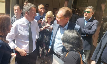 Two men in suits among a small cluster of people in the sunshine.