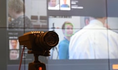 A mounted facial recognition camera with a screen in the background displaying images of people