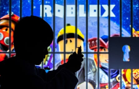 A boy poses for a photo while holding a smartphone in front of a screen displaying characters of the video game Roblox